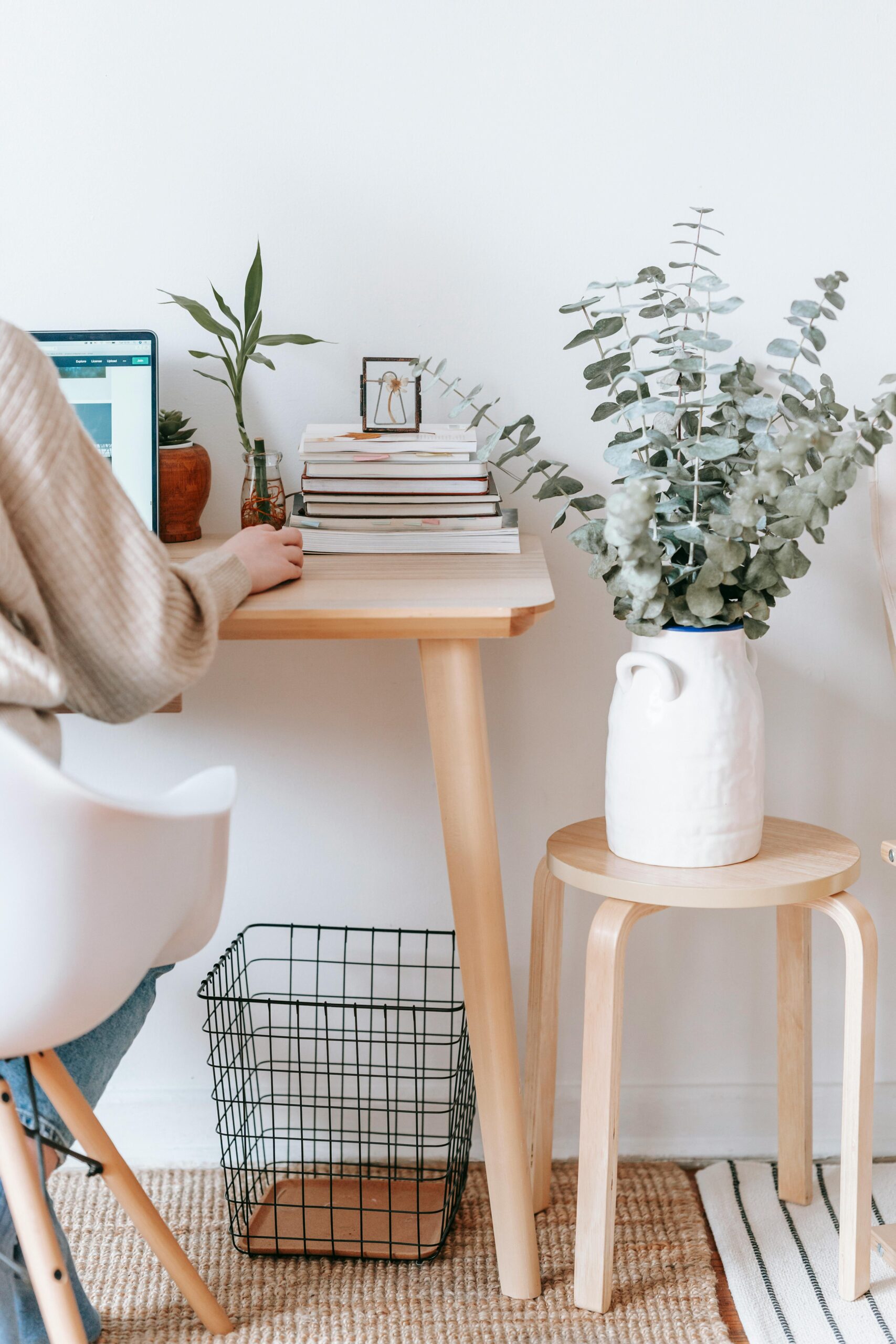 A cozy workspace with a wooden desk, a stack of books, plants, and a laptop on the left side.