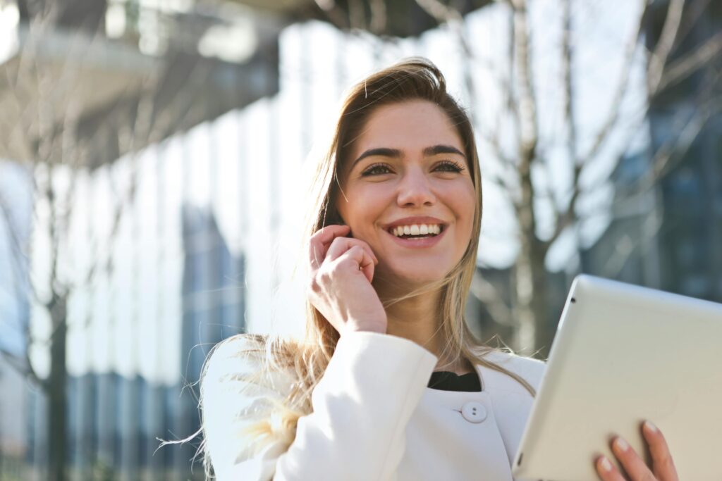 A smiling woman holding a tablet and talking on the phone outdoors with a blurred cityscape in the background.